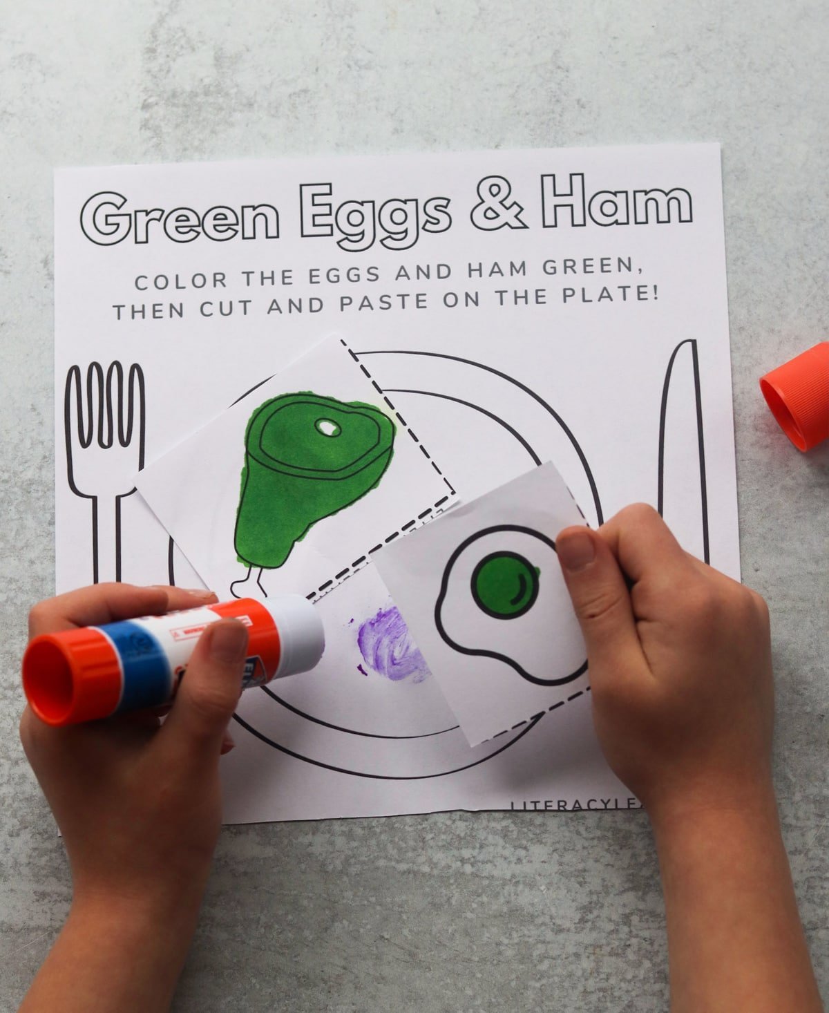 A child's hands glueing the cut pieces of the green eggs and ham activity page together. 