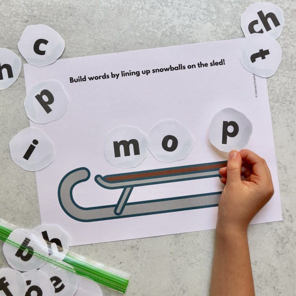 A child's hand arranging cut out snowball shaped letters on a sled worksheet.