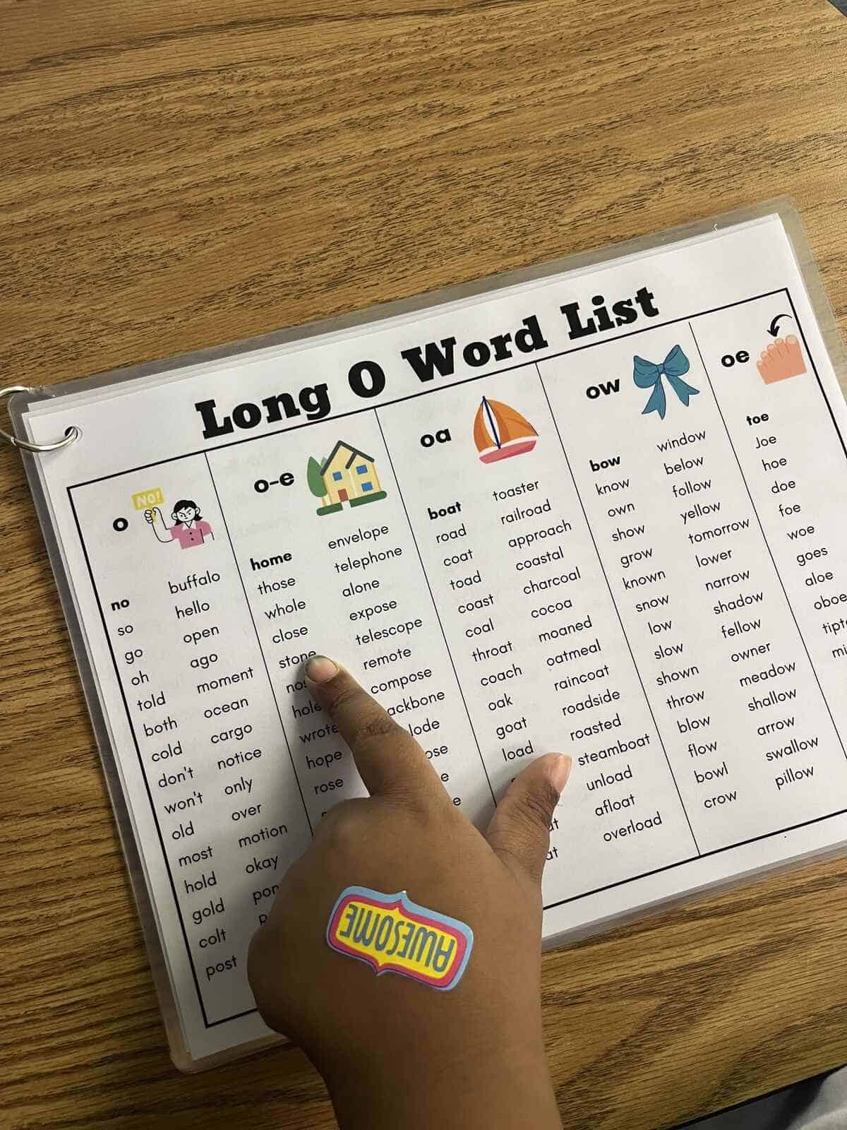 A child's hand pointing to a word on the Long O word list worksheet.