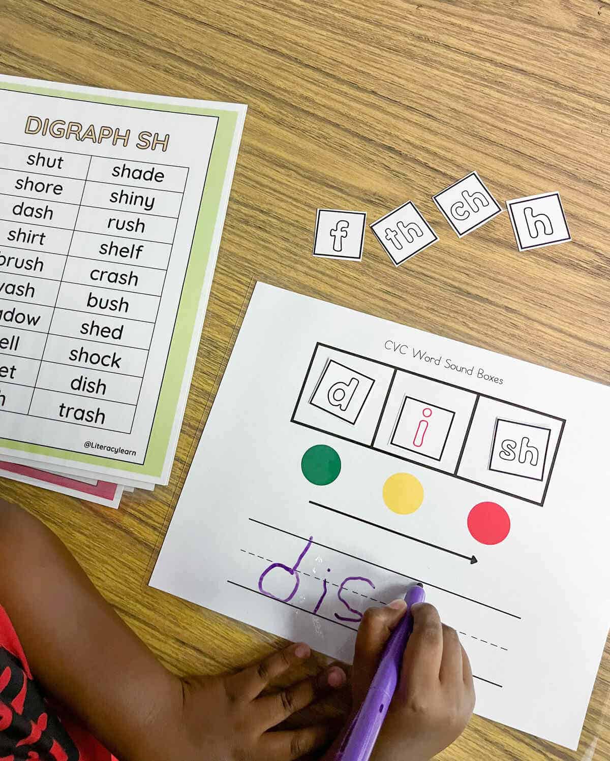 A child's hands writing a word on a worksheet with the digraph list beside it.