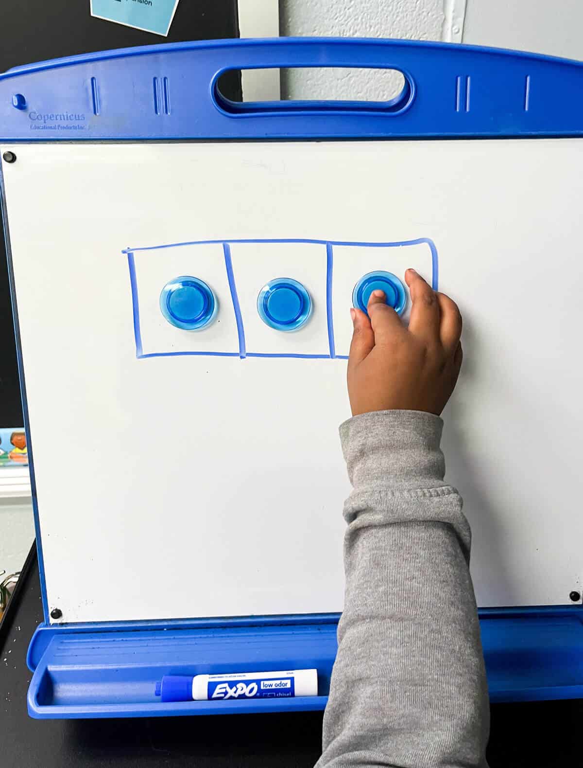 Hand-drawn Elkonin Boxes on a white board with a child's hand pushing blue markers.