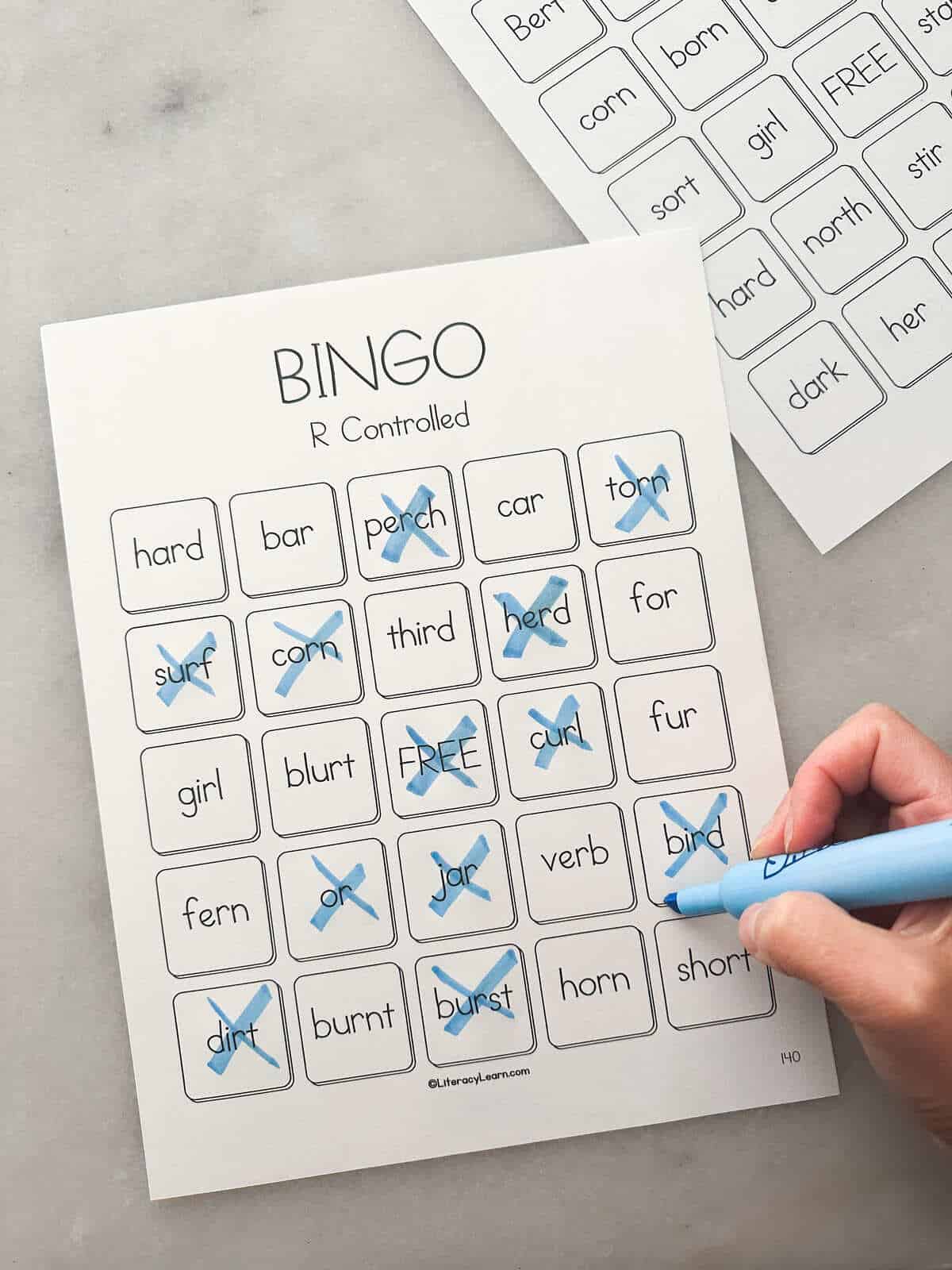 A child's hand marking words off with a blue marker on one R-Controlled Bingo card. 