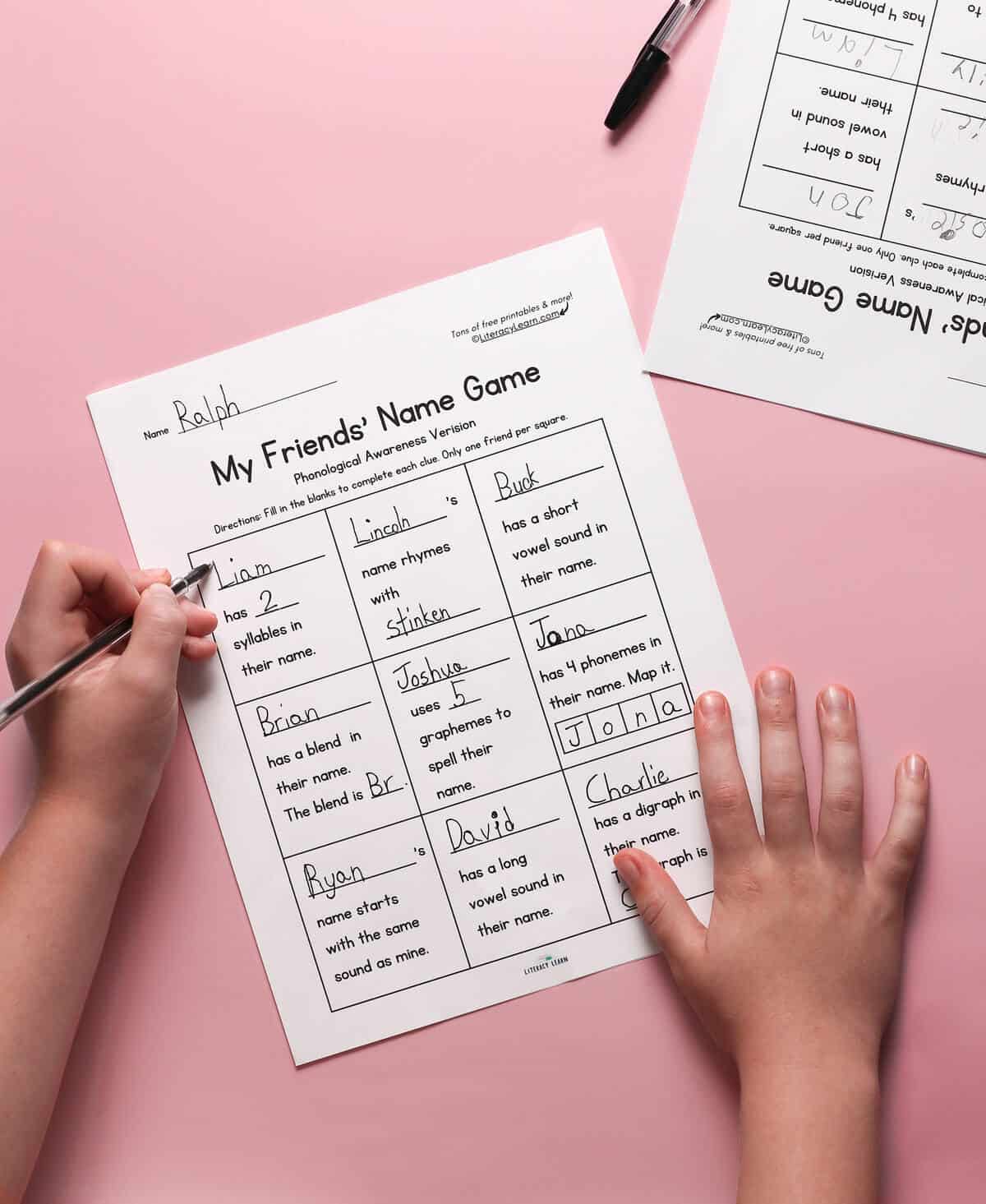 A child's hands holding a pen, filling out the Phonological Awareness Name Game worksheet.
