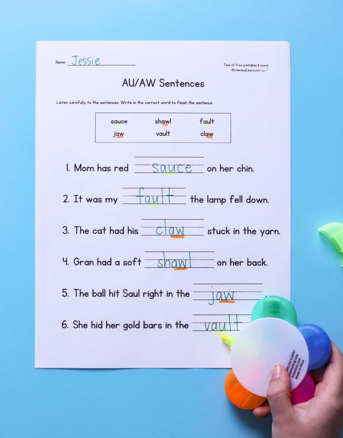 A child's hand with a printed and completed au/aw decodable sentences worksheet.
