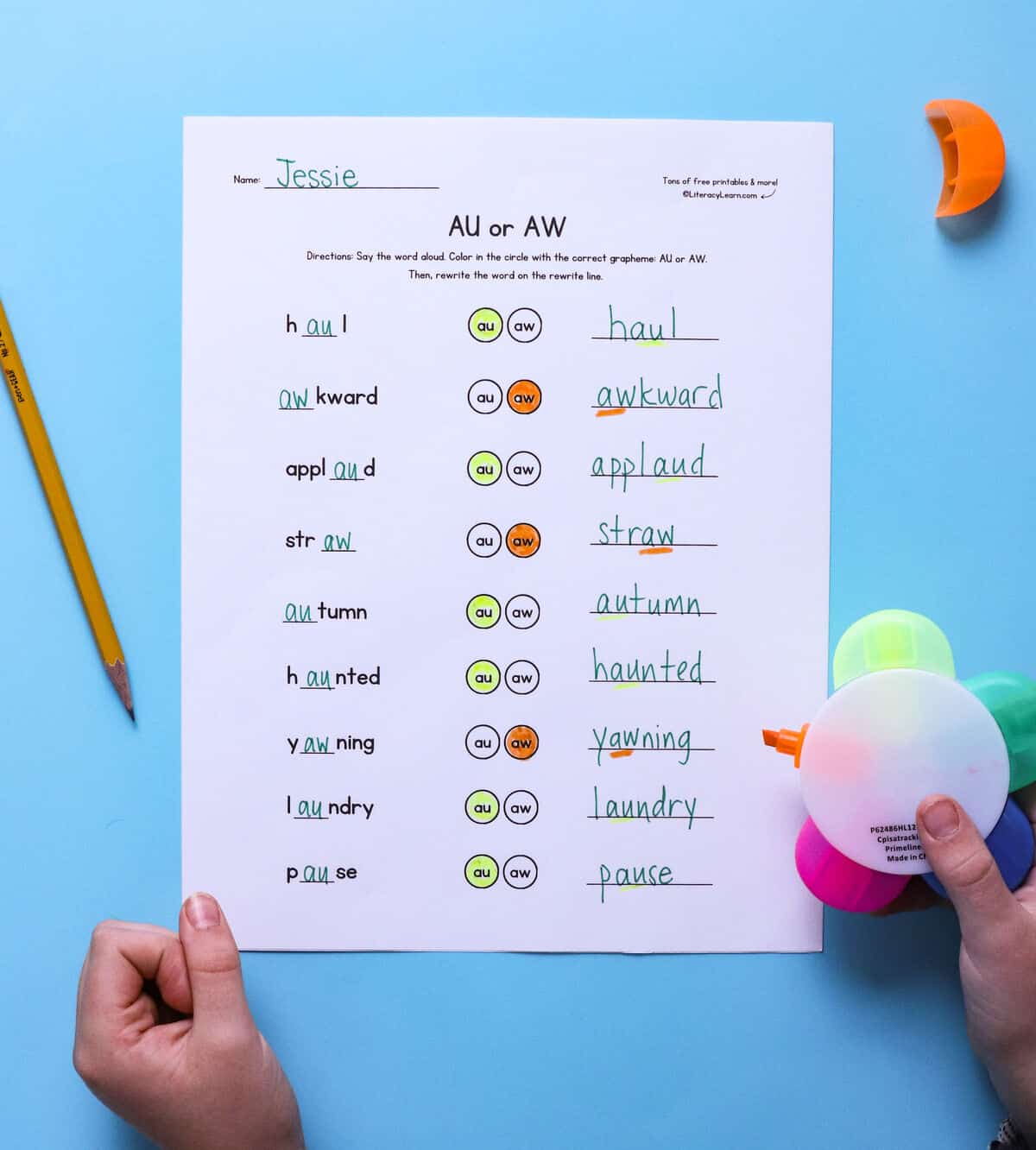 A child's hands highlighting words on a printed and completed au/aw spelling worksheet.