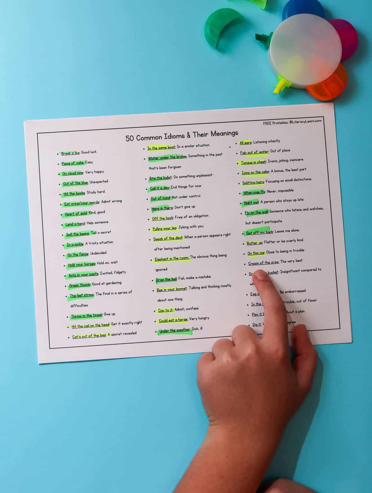 A child's hand pointing to a printed list of 50 common idioms.